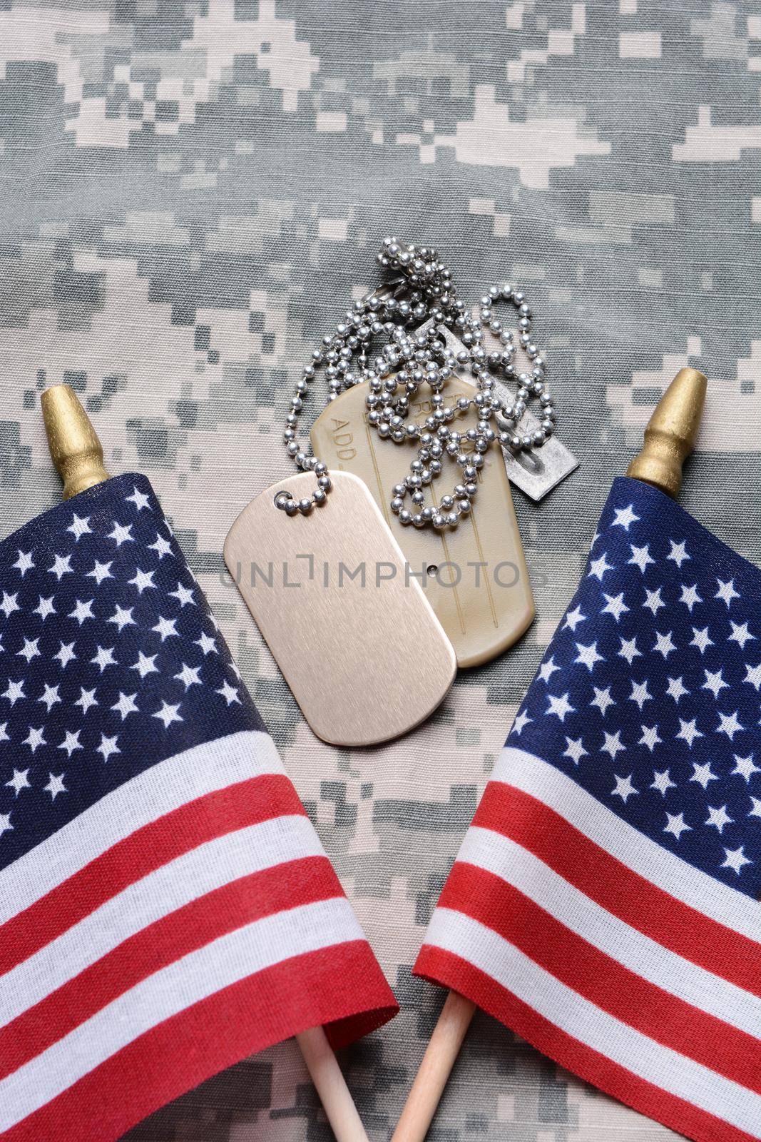 Closeup of two crossed American Flags on camouflage material with dog tags in the middle. The ID tags are blank. Vertical format filling the frame.