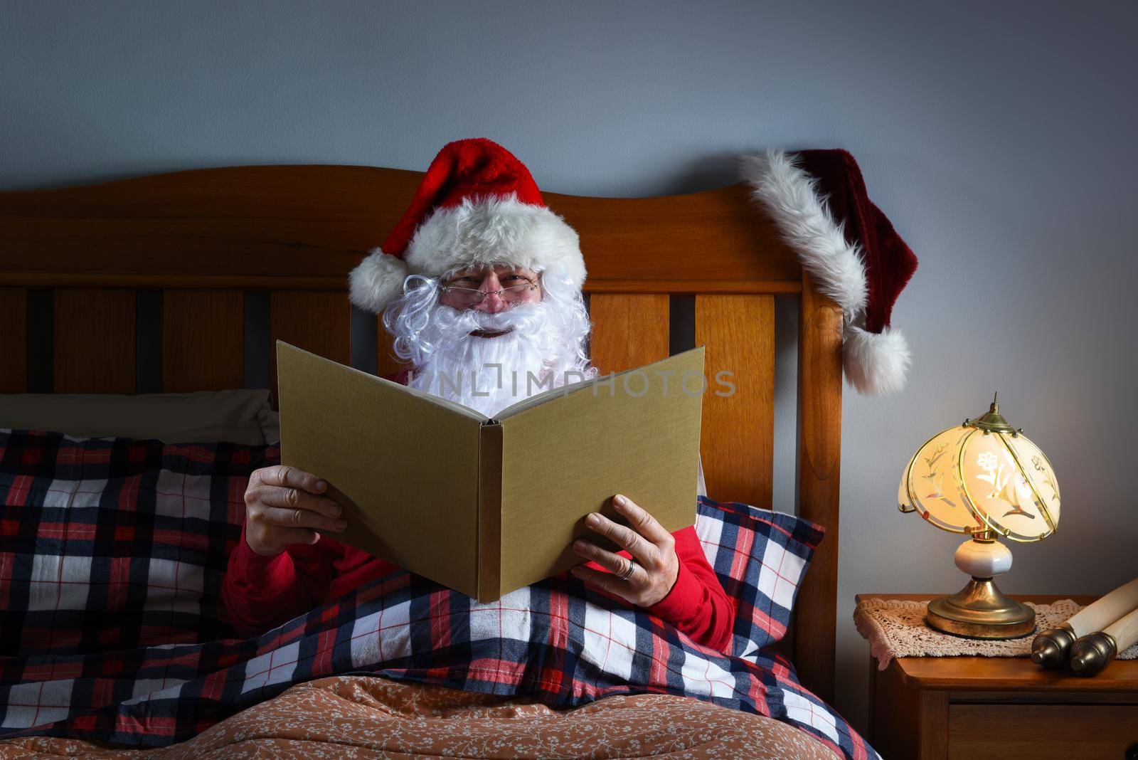 Santa Claus sitting in bed reading a large book before he goes to sleep on Christmas Eve.