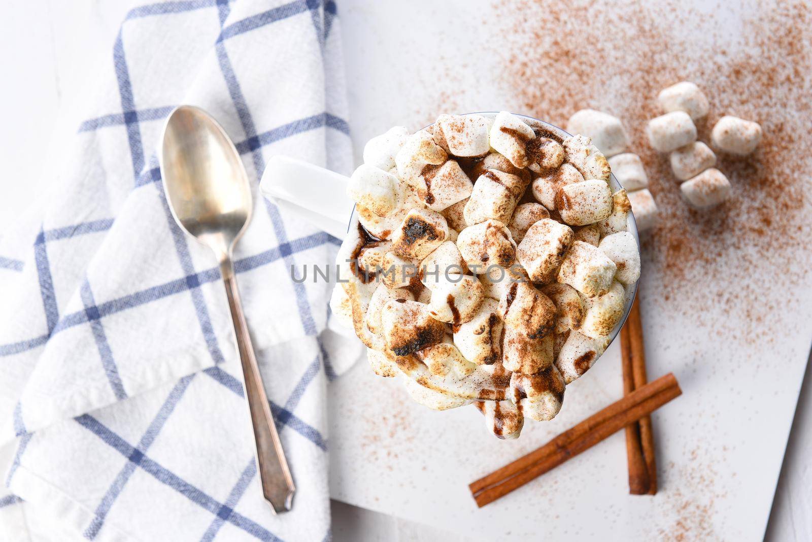 Overhead view of a mug of hot chocolate with toasted marshmallows sprinkled with cinnamon. A spoon towel and cinnamon sticks 