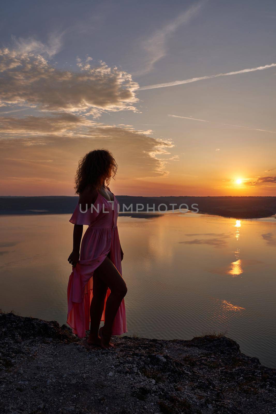 Seductive, charming girl with curly hair standing on shore of lake and holding end of long dress and showing leg. Beautiful model posing on background of sunset near lake.