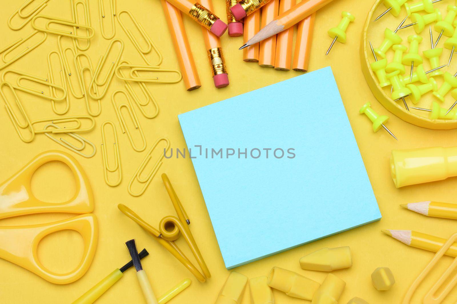 School supplies. Pencils, erasers, paper clips, brushes, pins, scissors, paper laying in a random pattern on a yellow background. Everything in a shade of yellow except for the blue note pad.