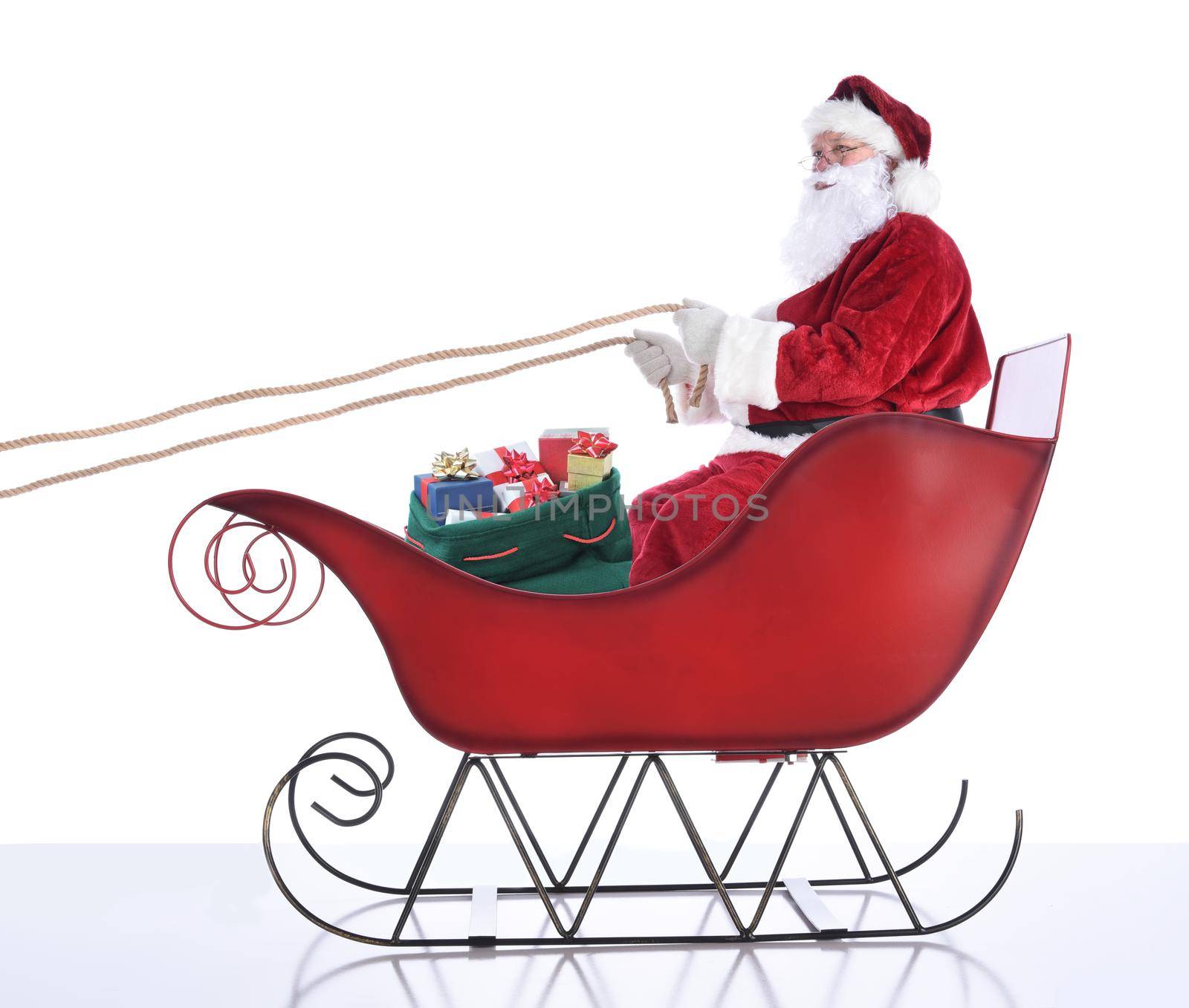 Santa Claus sitting in hi sleigh holidng onto the reins, with a sack of presents at his feet. Isolated on white.  by sCukrov
