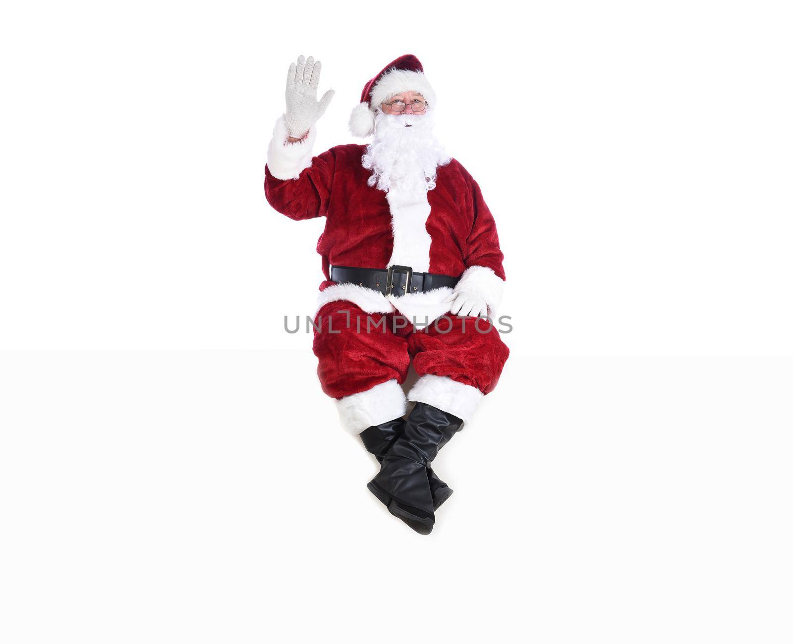 Senior man in traditional Santa Claus Suit sitting on a white wall waving.  Isolated on white with copy space. by sCukrov