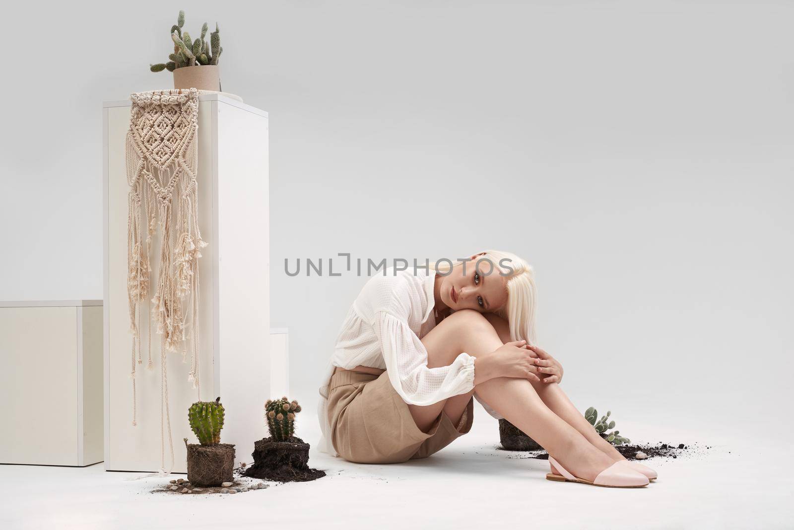 Charming blonde girl with beautiful make up in white blouse, beige shorts and ballet flats, sitting on floor of white studio with broken pots of green cactus. Concept of nature and beauty