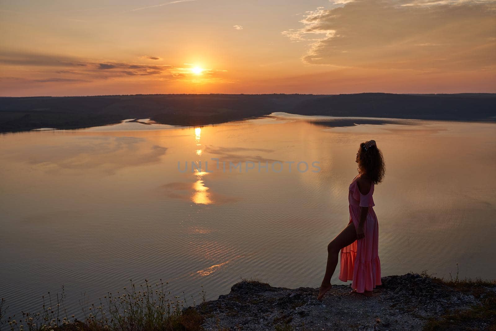 Beautiful model wearing in light silk dress posing on shore of lake. Girl looking at sun setting over horizon. Lady on background of sunset and water in evening.