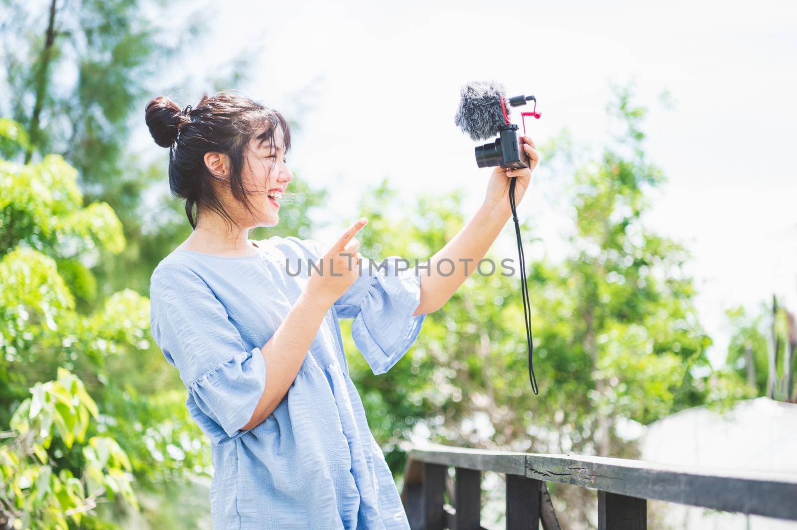 Asian woman in blue dress in public park carrying digital mirrorless camera and taking photo and vlog in happy mood. People lifestyle and leisure concept. Outdoor travel and Nature theme.