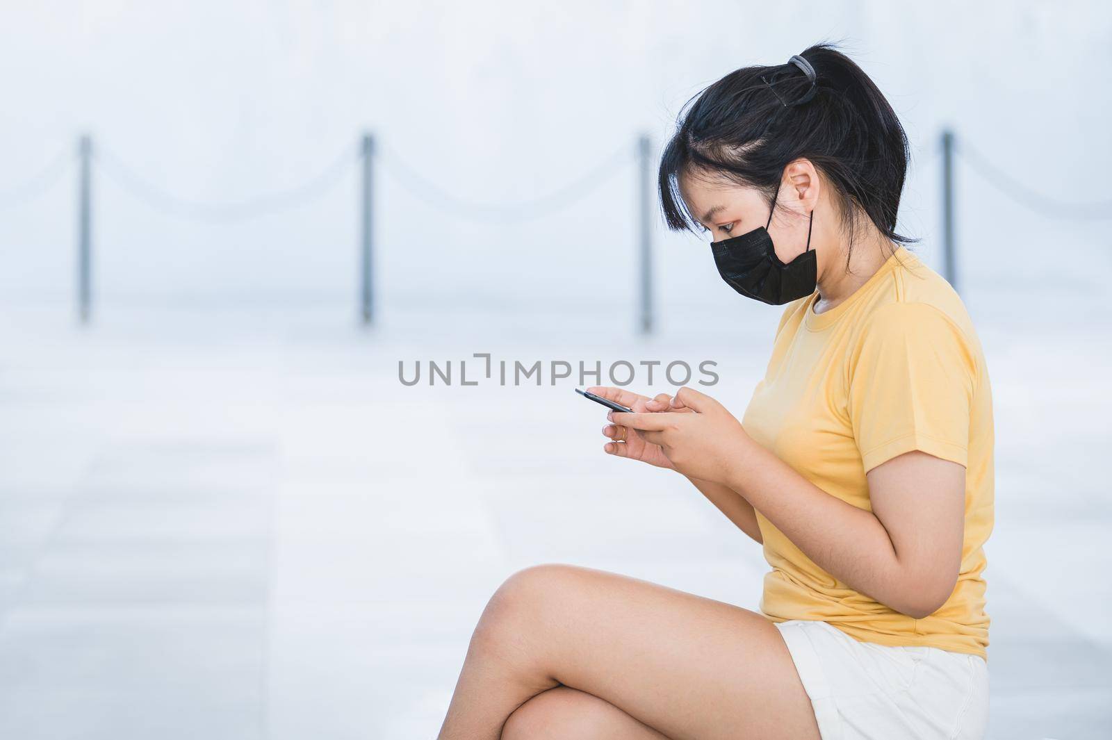 Asian woman wearing facial mask sits on and browsing social media to update COVID news via mobile phone in a new normal situation at outdoor. Technology and Medical concept by MiniStocker