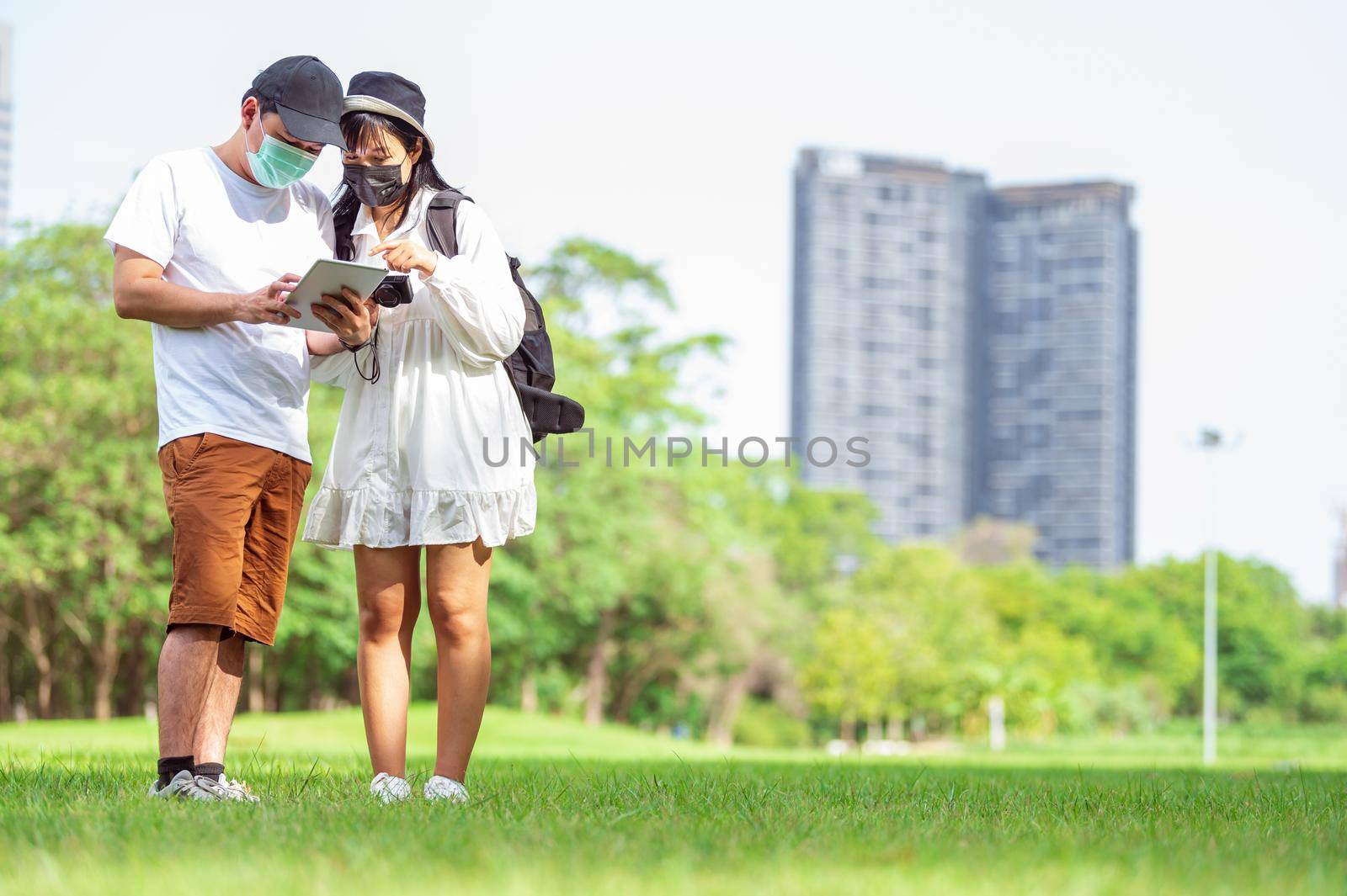 Asian couples with facial mask searching for tourist information through tablets of amazing places to visit in urban with building and park background. Technology and Travel concept. New normal theme