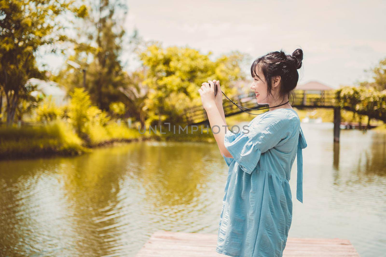 Asian woman in blue dress in public park carrying digital mirrorless camera and taking photo without facial mask in happy mood. People lifestyle and leisure concept. Outdoor travel and Nature theme.