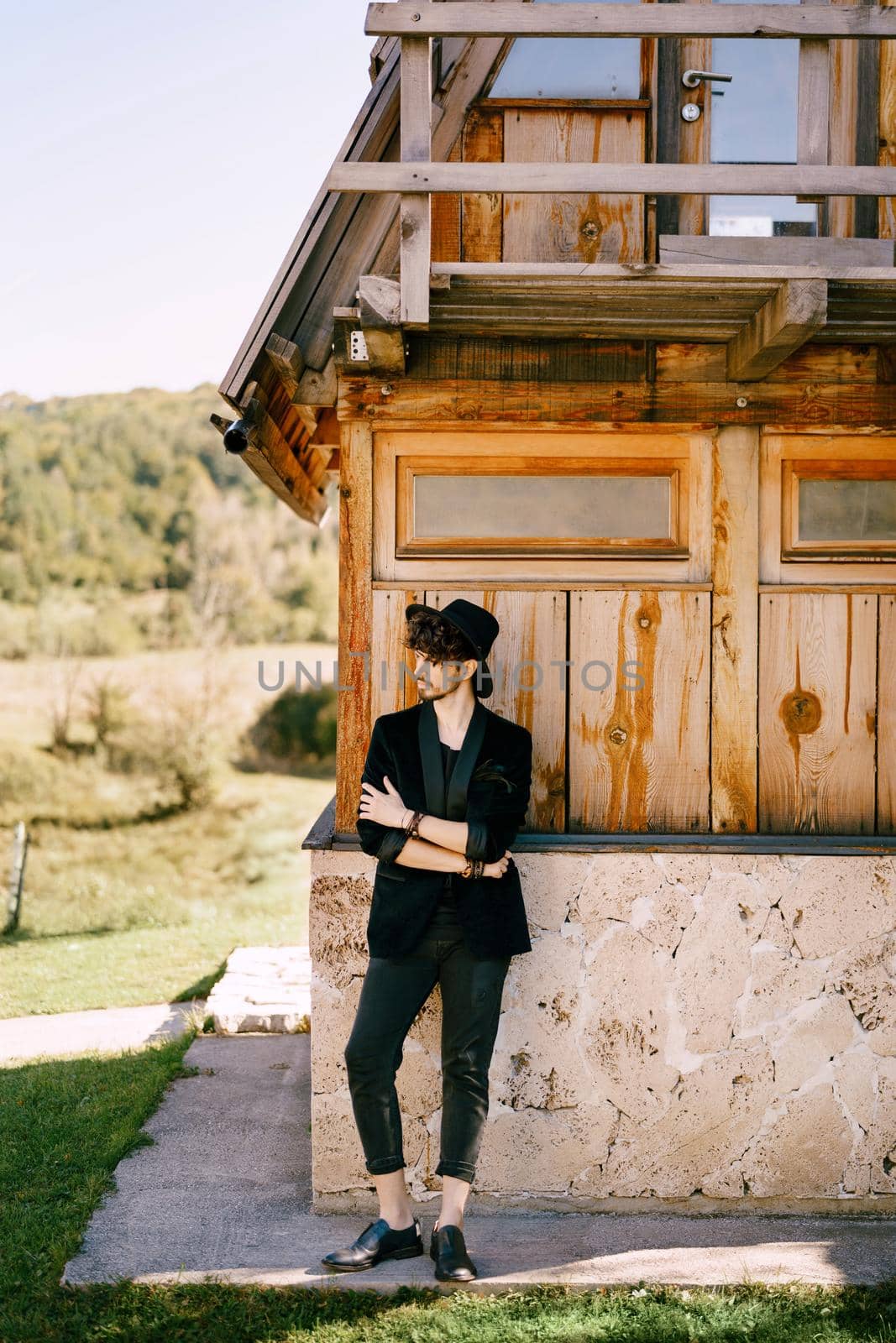 Young man in a hat stands near an old wooden house with his arms folded on his chest by Nadtochiy