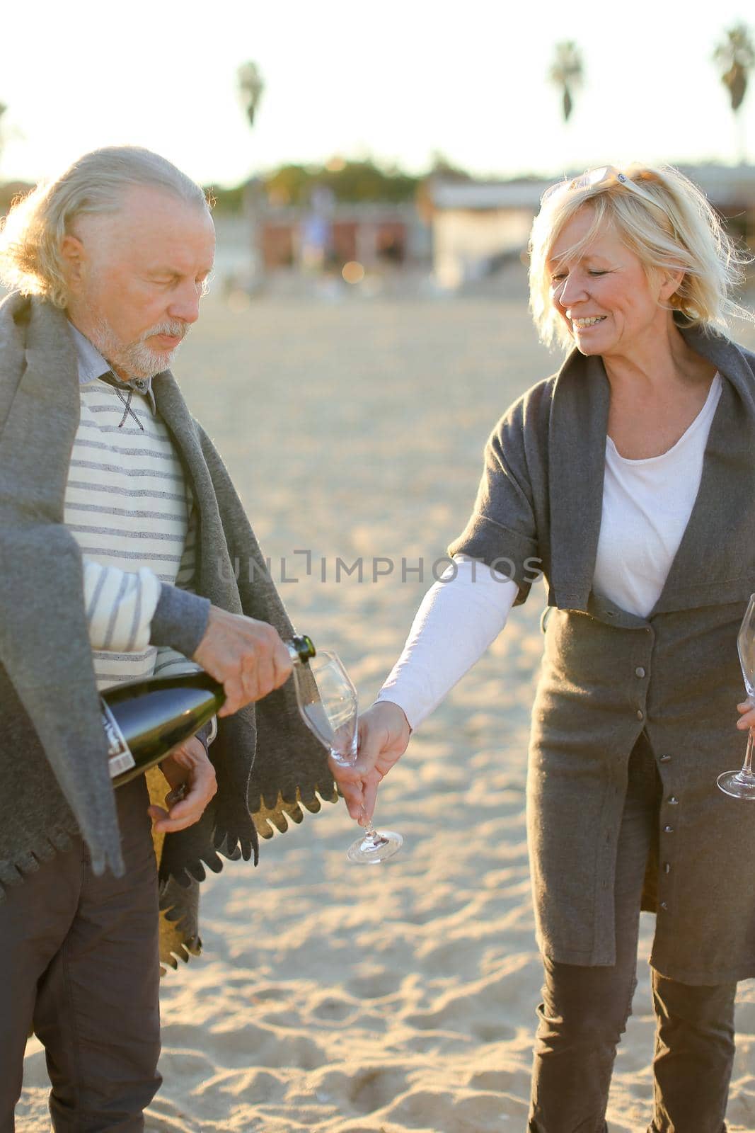 Focus on pouring champagne by elderly husband with caucasian wife on sand beach. Concept of picnic in open air.