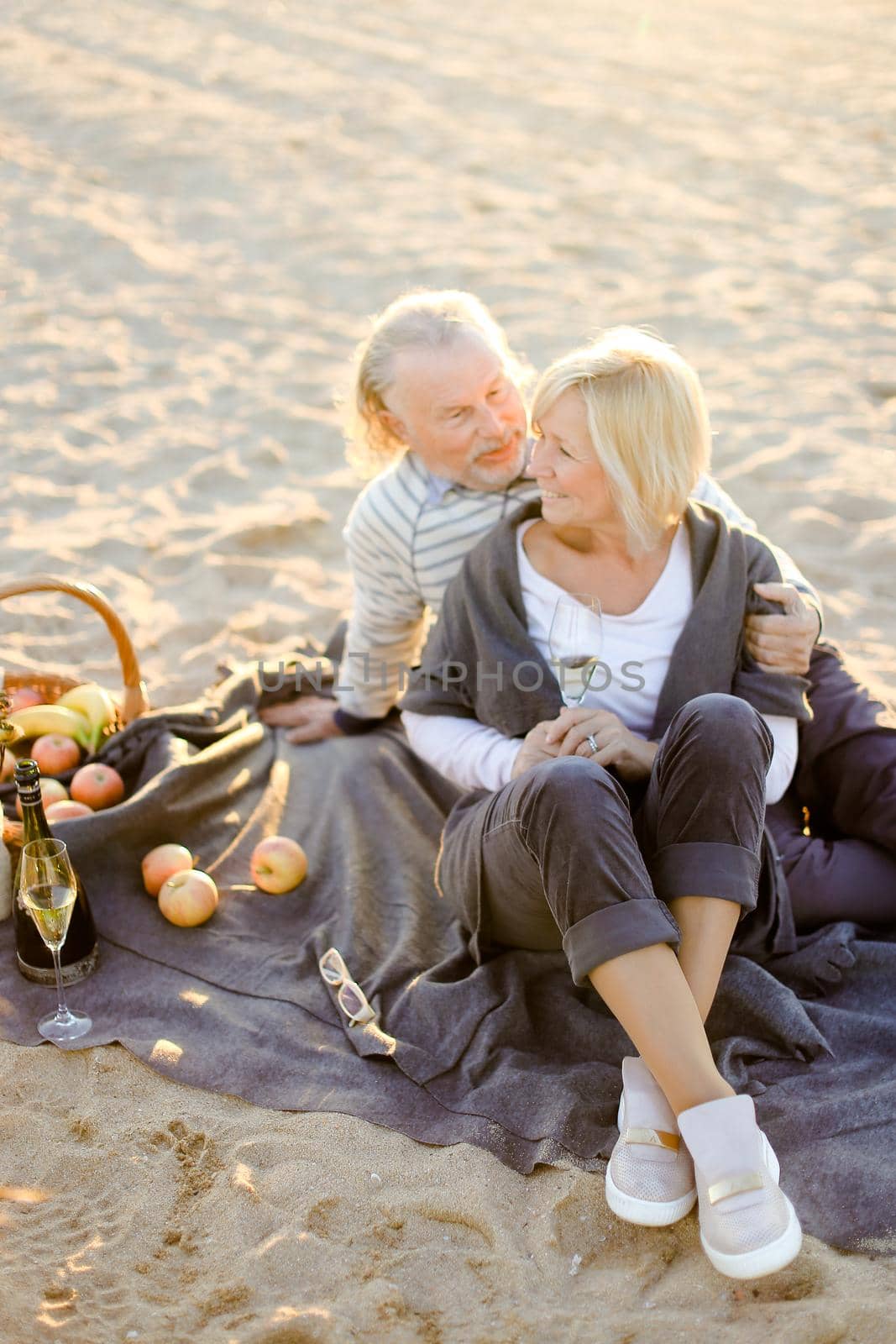 Senior husband hugging elderly wife sitting on plaid with champagne on sand beach. by sisterspro