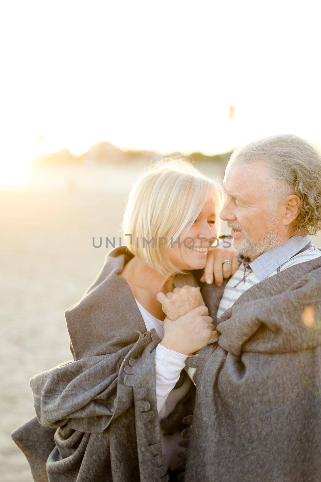 Sunshine photo of senior husband hugging blonde wife wearing plaid on sand beach. Concept of happy elderly couple and relationship.