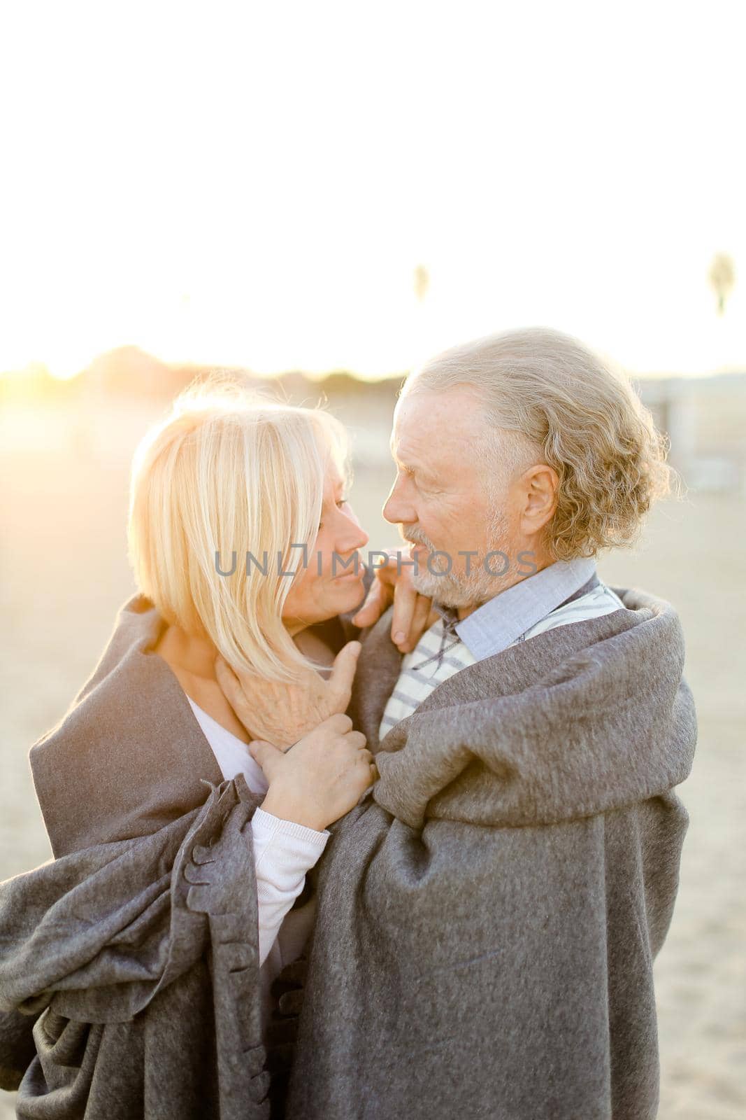 Sunshine photo of caucasian senior husband hugging wife wearing plaid on sand beach. by sisterspro