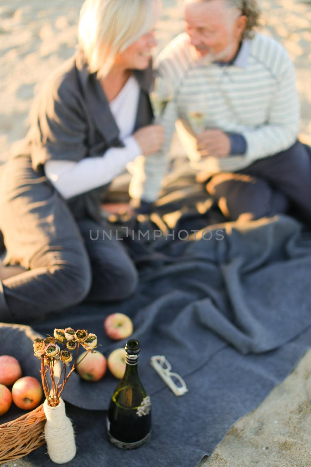 Focus on champagne bottle and fruits, pensioners with glasses sitting on plaid on sand beach in blurry background. Concept of elderly couple on picnic.