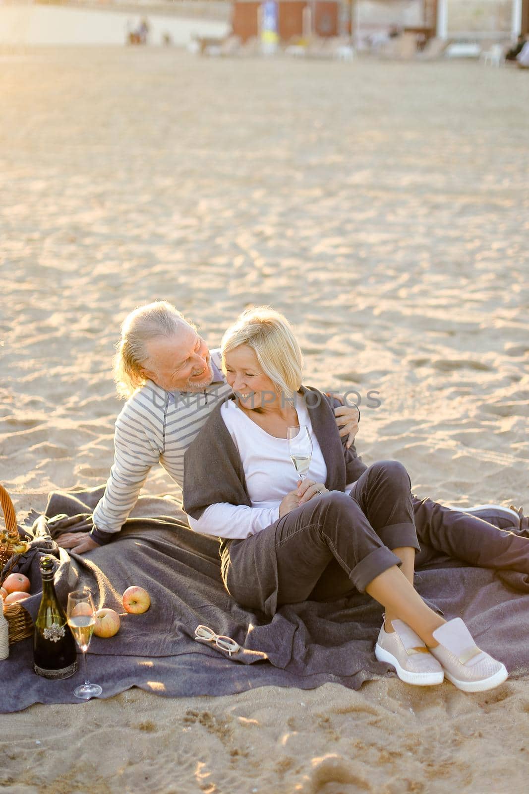 Sunshine photo of senior husband hugging wife sitting on plaid with champagne on sand beach. by sisterspro