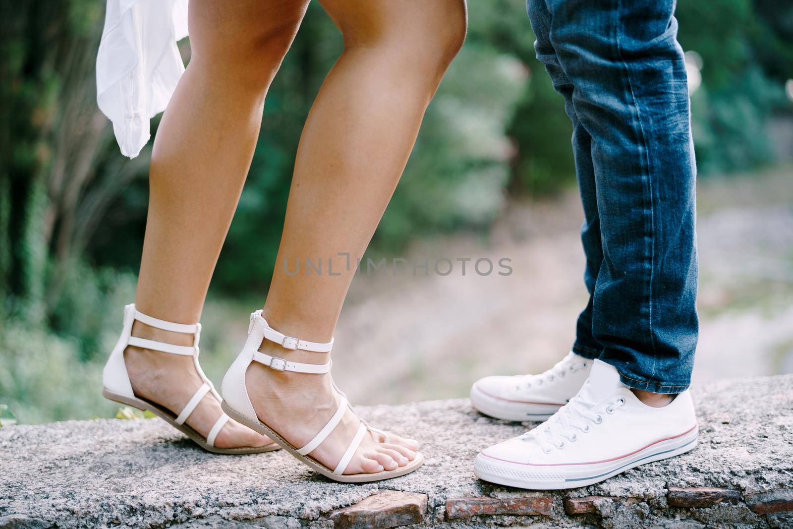 Female feet in sandals next to male feet in sneakers stand on a stone border in the park by Nadtochiy