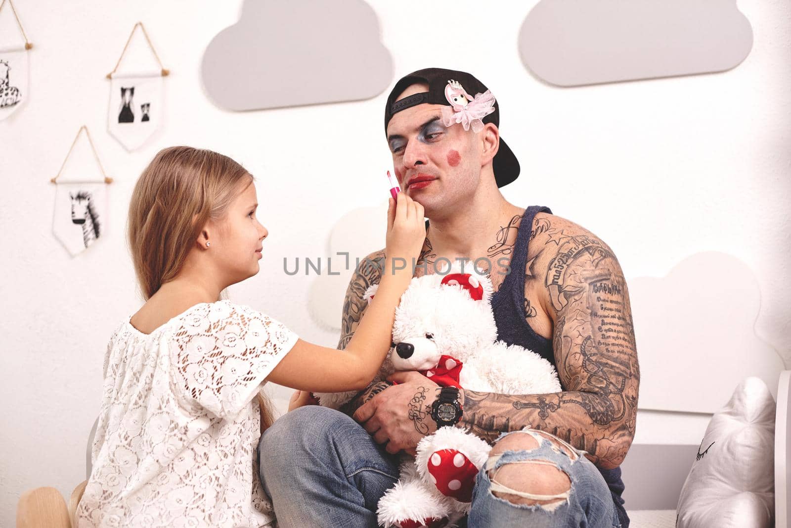 Funny time Tattoed father in a cap and his child are playing at home. Pretty girl in a white dress is putting a rouge at her dady's cheeks while he is holding a toy bear, in her bedroom. Family holiday and togetherness.