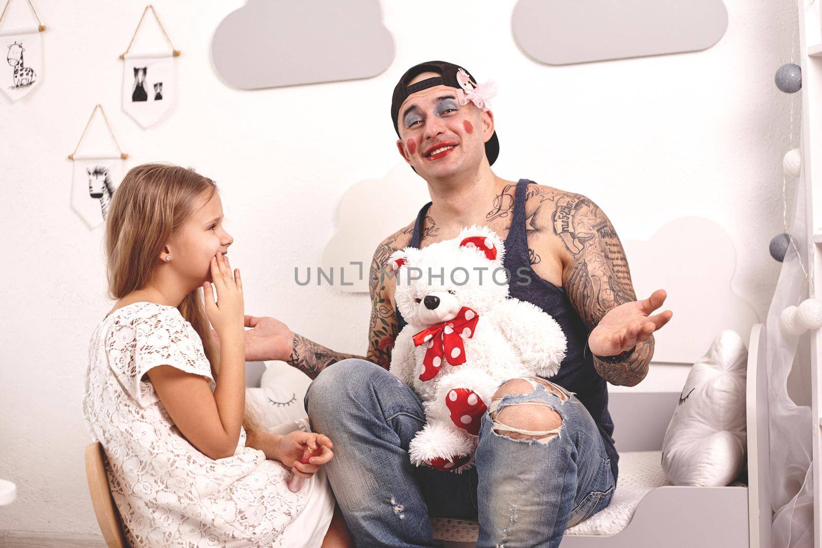 Funny time Tattoed father in a cap, whith a toy bear in his hands, and his child are playing at home. Pretty girl in a white dress is doing makeup to her dad and laughing at him, in her bedroom. Family holiday and togetherness.