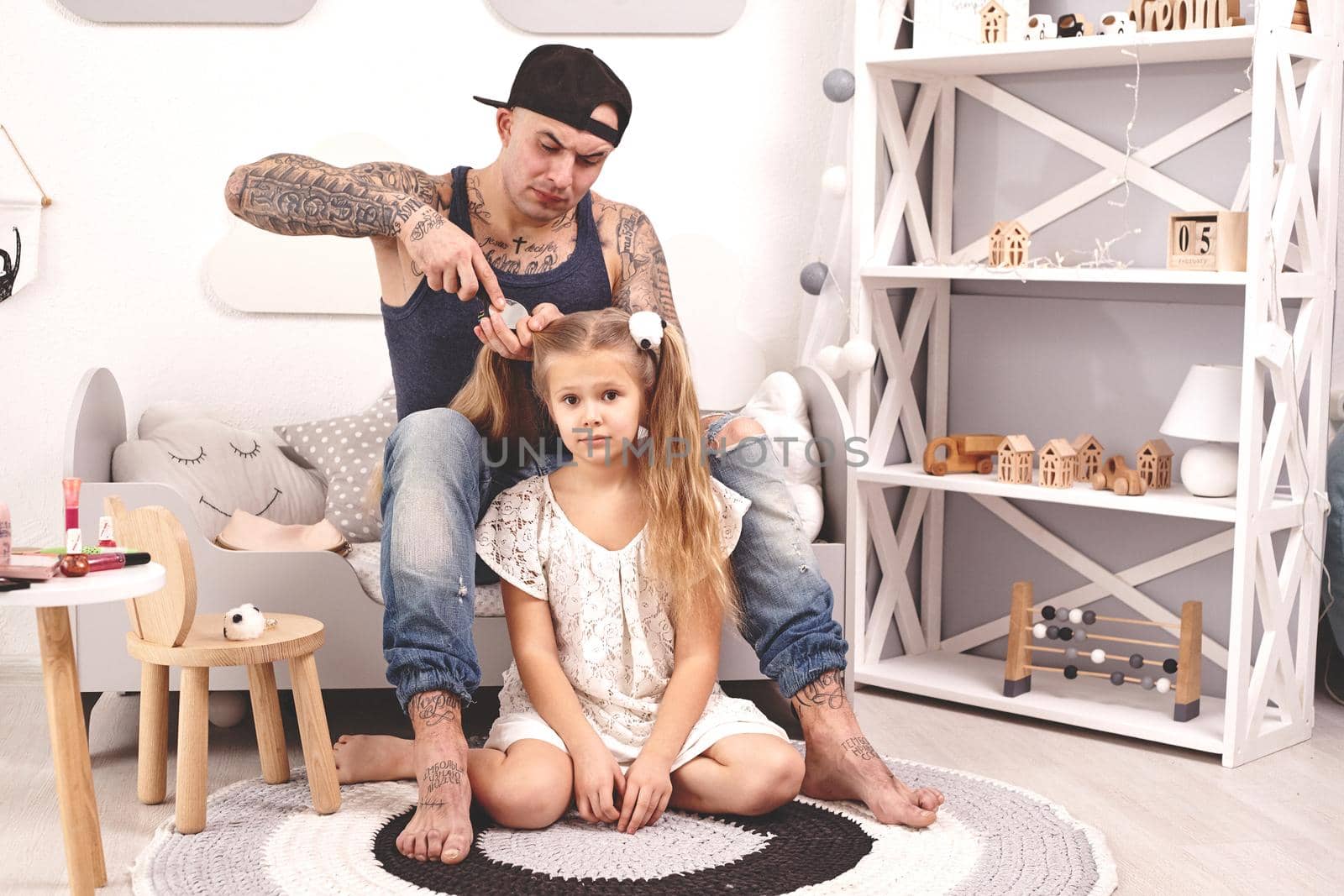 Funny time Attractive young father in a cap and his child are playing at home. Dad is doing his daughter's hair while she is sitting on a rug in her bedroom. Family holiday and togetherness.