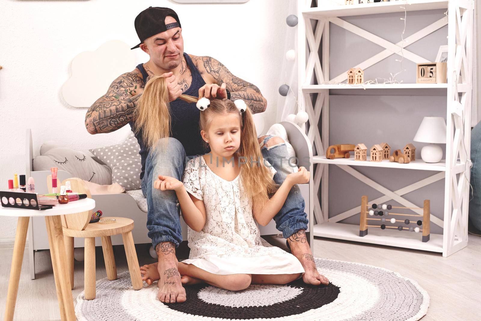 Funny time Tattoed father in a cap and his child are playing at home. Dad is doing his daughter's hair in her bedroom. Family holiday and togetherness by nazarovsergey