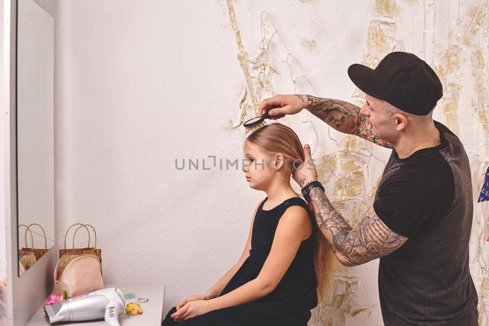 Cute little daughter and her tattoed dad are playing together near a mirror. Handsome dad is doing his daughter's hair. Family holiday and togetherness.