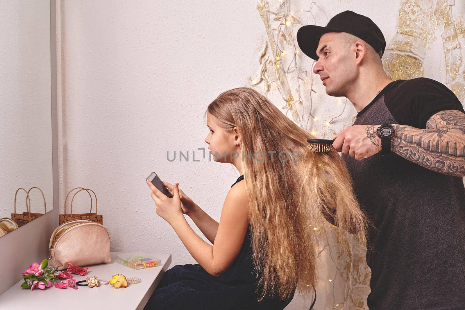 Cute little daughter and her tattoed dad are playing together near a mirror. Handsome dad is drying his daughter's hair while she is looking at her phone. Family holiday and togetherness.