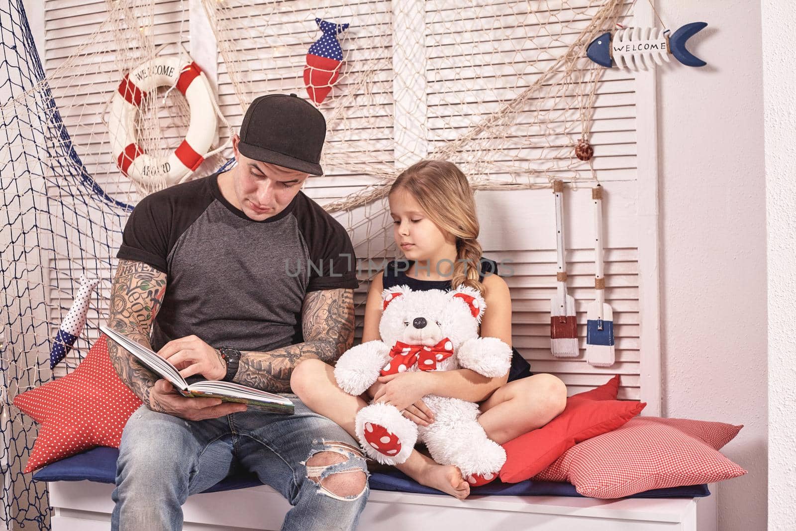 Young tattoed man in a cap is spending time with his little cute daughter. They are sitting on a white bench with some red pillows, in a room decorated in a marine style. She is holding a toy bear. Reading fairytales while daughter is sitting nearby. Happy family.