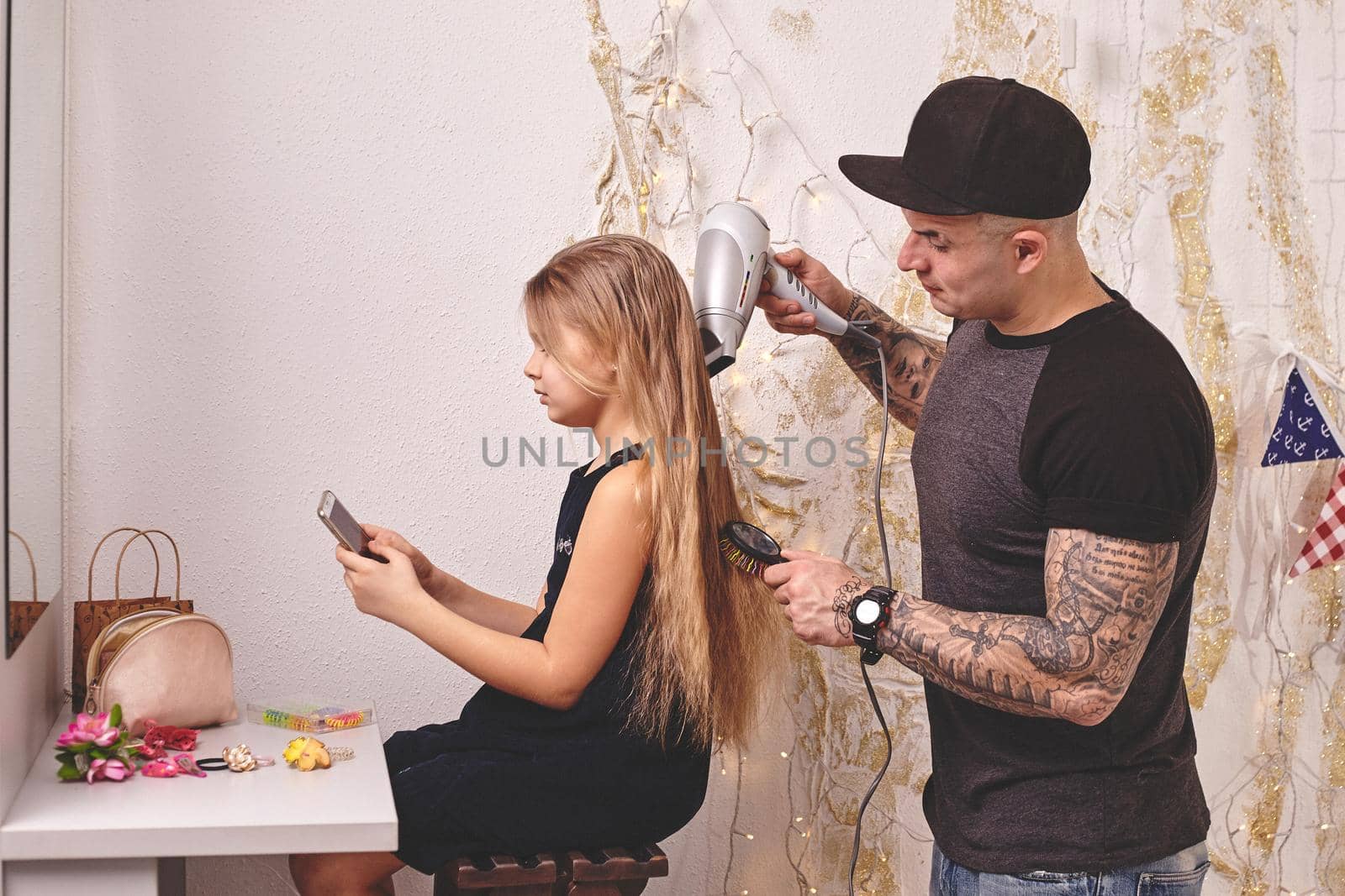 Cute little daughter and her tattoed dad are playing together near a mirror. Dad is drying his daughter's hair while she is looking at her phone. Family holiday and togetherness.