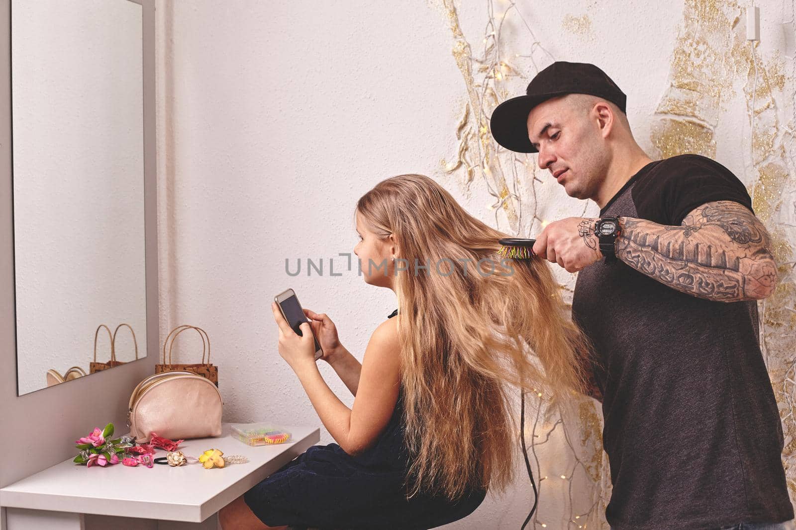 Cute little daughter and her tattoed dad are playing together near a mirror. Young dad is drying his daughter's hair while she is looking at her phone. Family holiday and togetherness.