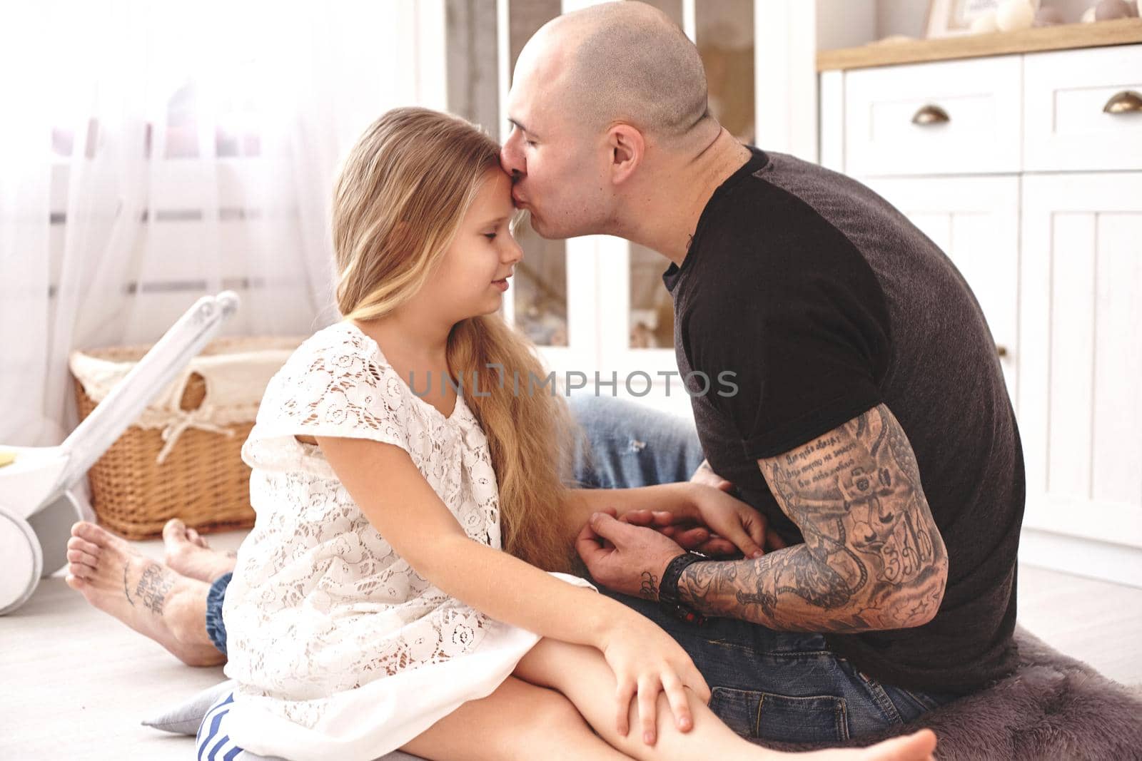 White modern kid's room whith a wooden furniture. Adorable daughter with a blond long hair wearing a white dress. Daddy with tattoos is kissing her while holding her hand. Friendly family spending their free time together sitting on a pillows.