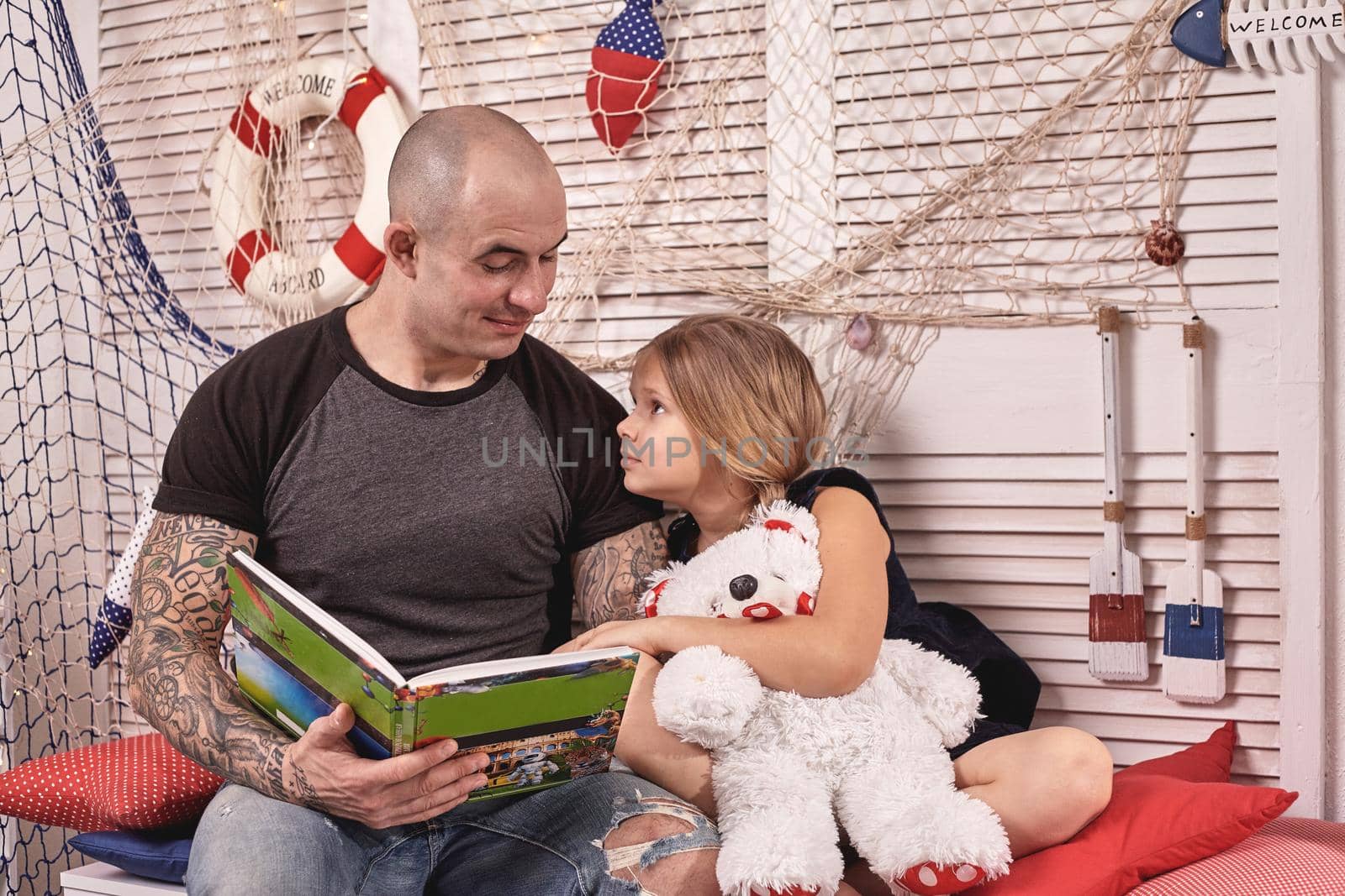 Bald tattoed man in a cap is spending time with his little cute daughter. They are sitting on a white bench with some red pillows, in a room decorated in a marine style. She is holding a toy bear and they are looking at each other. Reading fairytales while daughter is sitting nearby. Happy family.