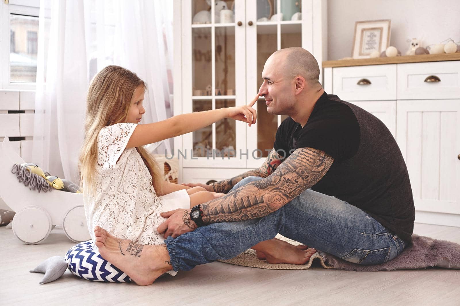 White modern kid's room whith a wooden furniture. Adorable daughter wearing a white dress is touching daddy's nose and looking with tenderness at him. Daddy with tattoos is hugging her. Friendly family spending their free time together sitting on a pillows.