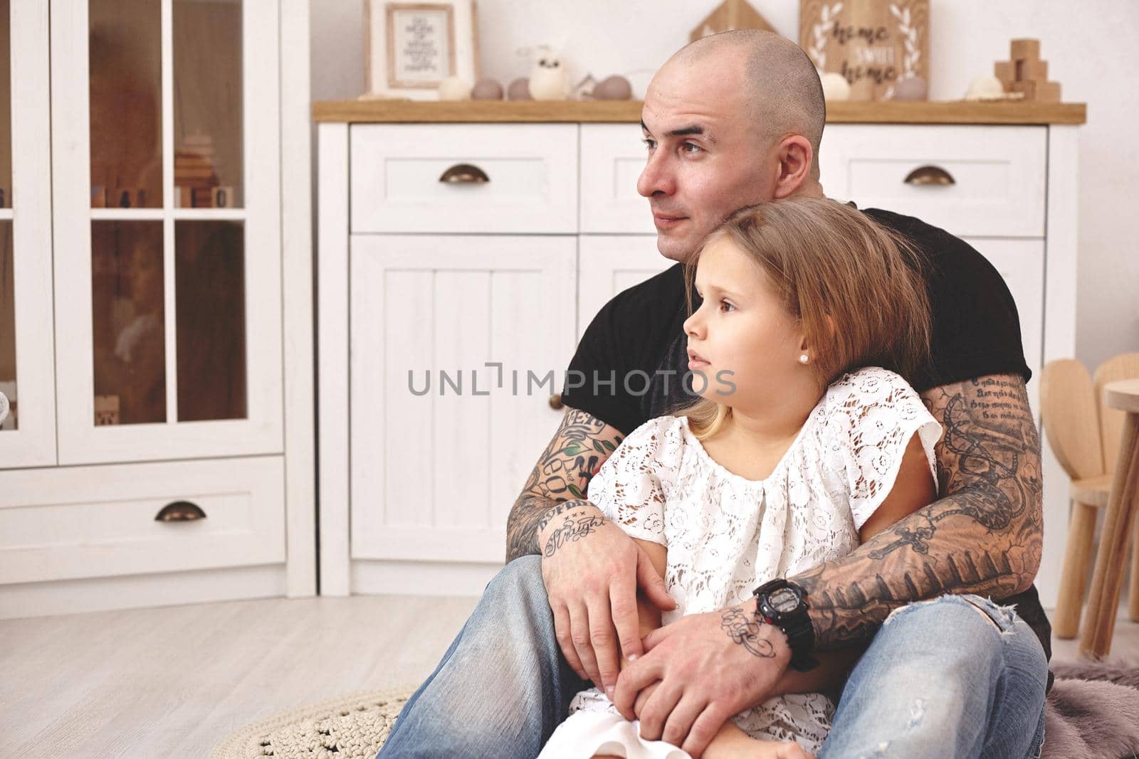 White modern kid's room whith a wooden furniture. Adorable daughter with a long blond hair wearing a white dress. Daddy with tattoos is hugging her and they are looking somewhere. Friendly family spending their free time together sitting on a pillows.