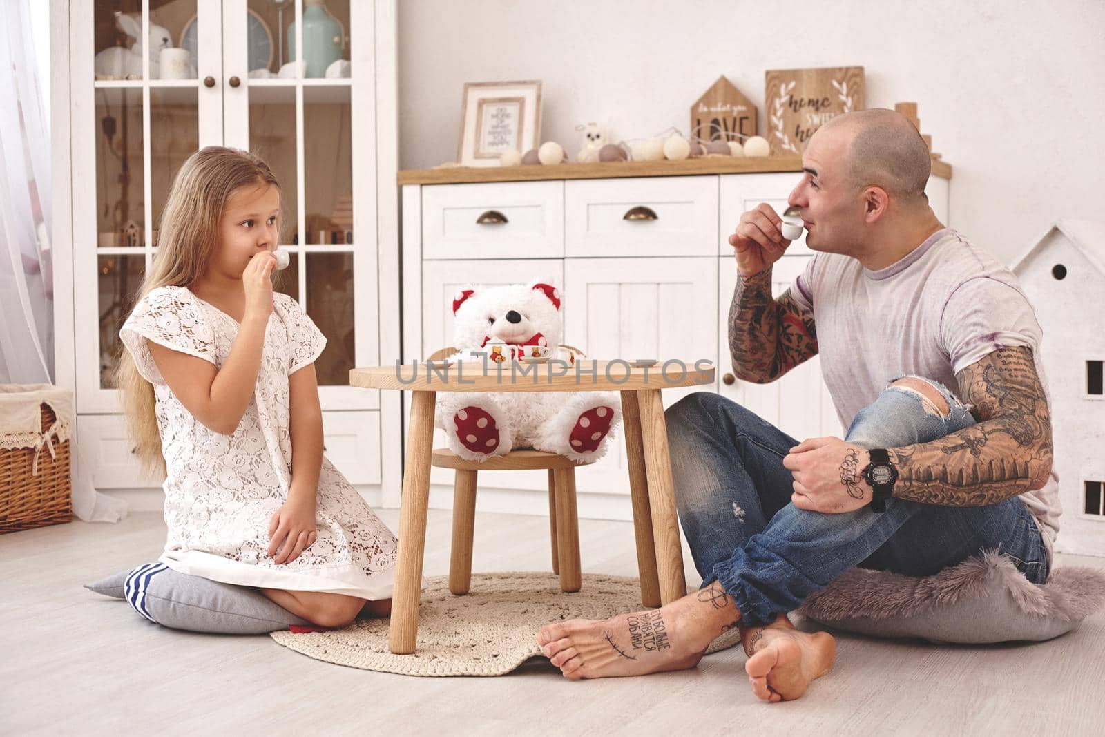 White modern kid's room whith a wooden furniture. Adorable daughter wearing a white dress is looking with tenderness at her loving father. They are drinking tea from a toy dishes in a modern kid's room whith a wooden furniture. Daddy with tattoos is looking at her. Friendly family spending their free time together sitting on a pillows.