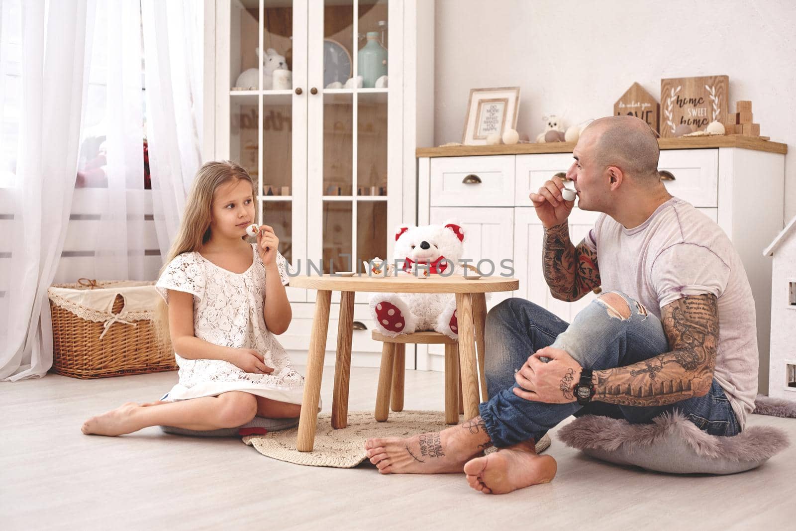 White modern kid's room whith a wooden furniture. Adorable daughter wearing a white dress is looking with tenderness at her loving father. They are drinking tea from a toy dishes in a modern kid's room whith a wooden furniture. Young daddy with tattoos is looking at her. Friendly family spending their free time together sitting on a pillows.