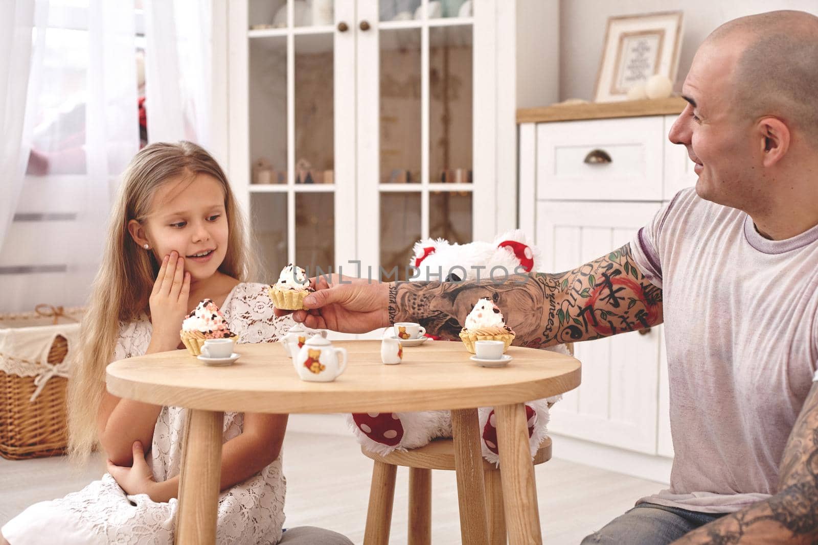 White modern kid's room whith a wooden furniture. Adorable daughter wearing a white dress with a long blond hair. They are drinking tea from a toy dishes in a modern kid's room whith a wooden furniture. Daddy with tattoos is treating his daughter with a cupcacke. Friendly family spending their free time together sitting on a pillows.