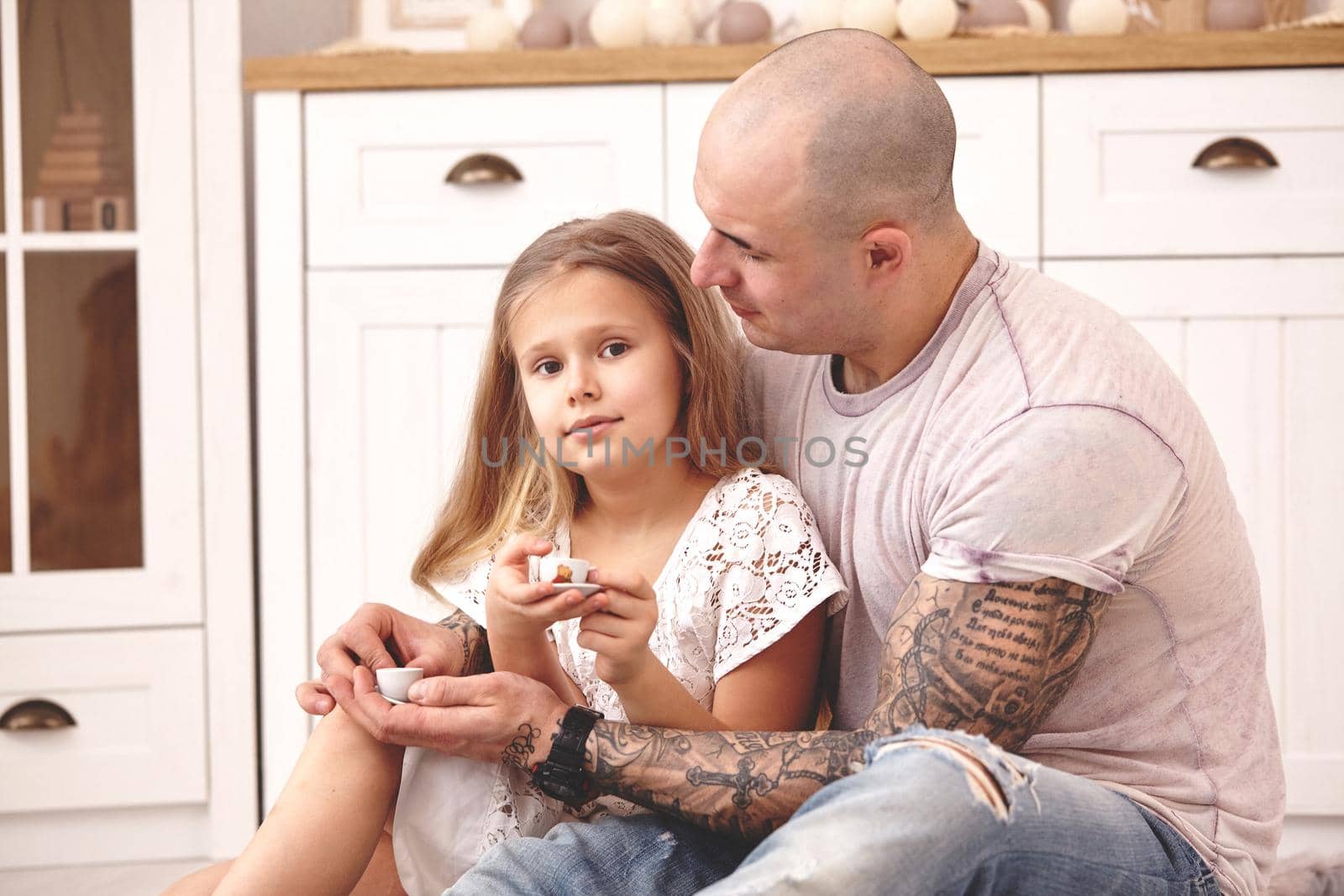 White modern kid's room whith a wooden furniture. Adorable daughter whith a long blond hair wearing a white dress is looking at the camera. They are drinking tea from a toy dishes in a modern kid's room whith a wooden furniture. Daddy with tattoos is hugging and looking at her. Friendly family spending their free time together sitting on a pillows.