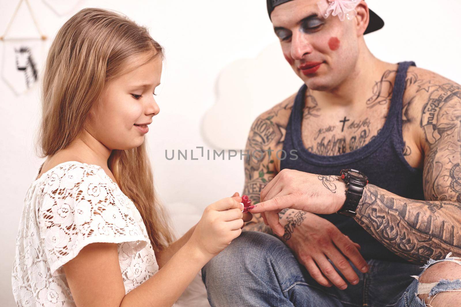 Funny time Tattoed father in a cap and his child are playing at home. Cute girl is doing makeup to her dad in her bedroom. Family holiday and togetherness by nazarovsergey