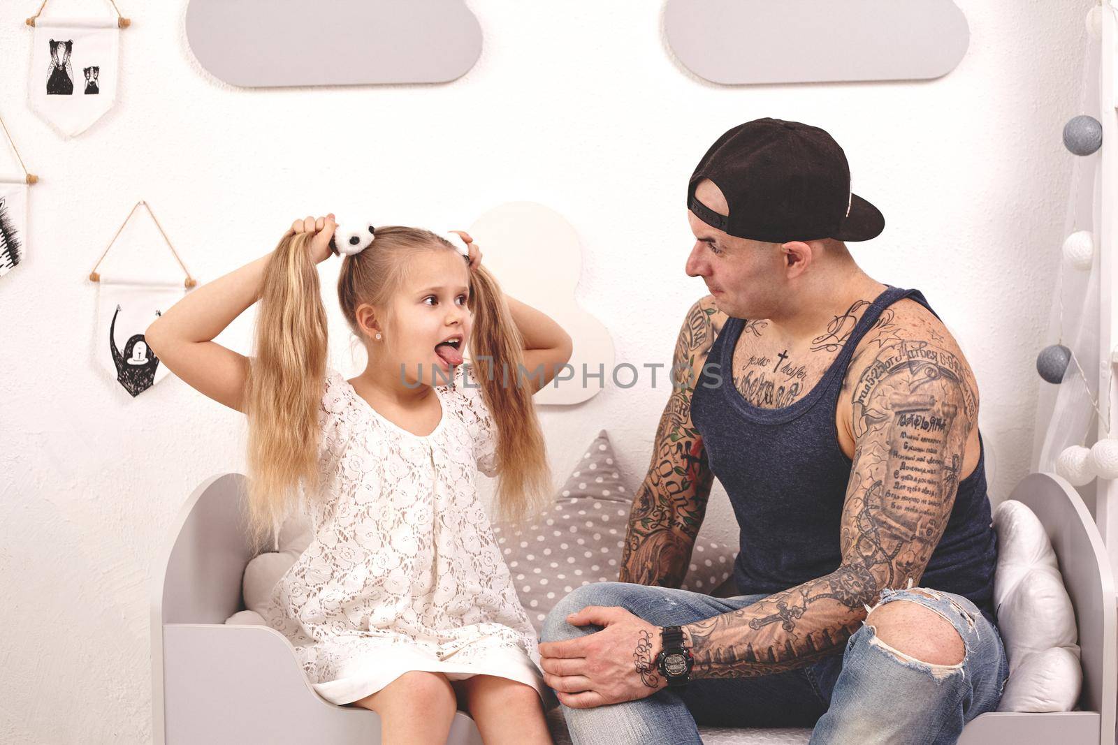 Funny time Tattoed father in a cap and his child are playing at home. Dad is doing his daughter's hair in her bedroom. They are looking at each other and she is showing her tongue. Family holiday and togetherness.