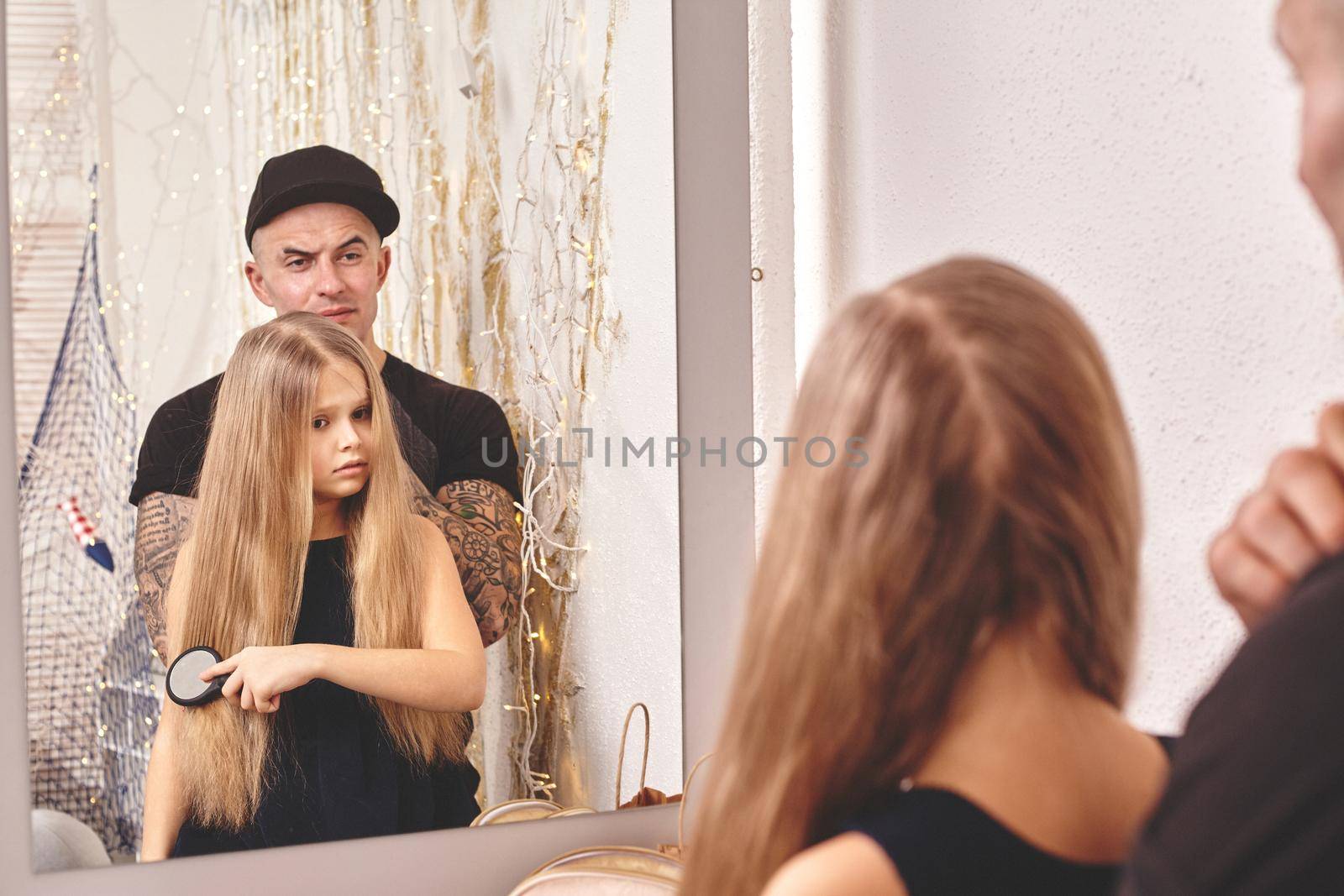 Cute little daughter and her tattoed dad are playing together near a mirror. Girl is combing her hair and daddy is looking at her. Family holiday and togetherness.