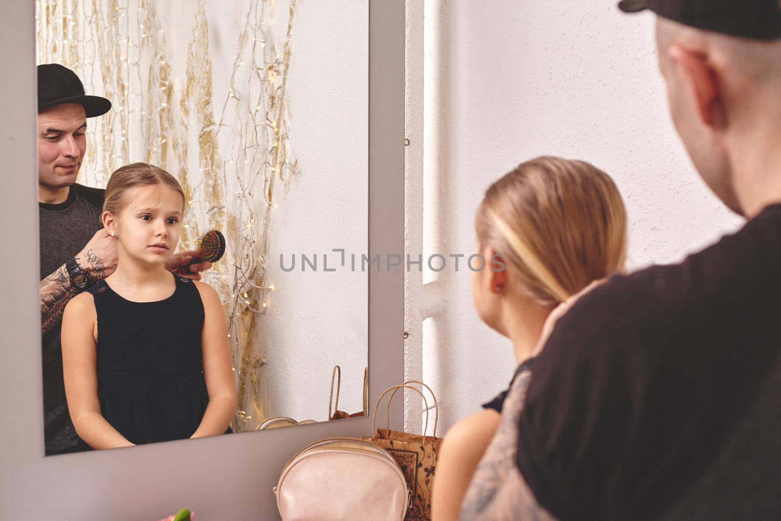 Cute little daughter and her tattoed dad are playing together near a mirror. Dad is doing his daughter's hair. Girl is looking at the mirror. Family holiday and togetherness.