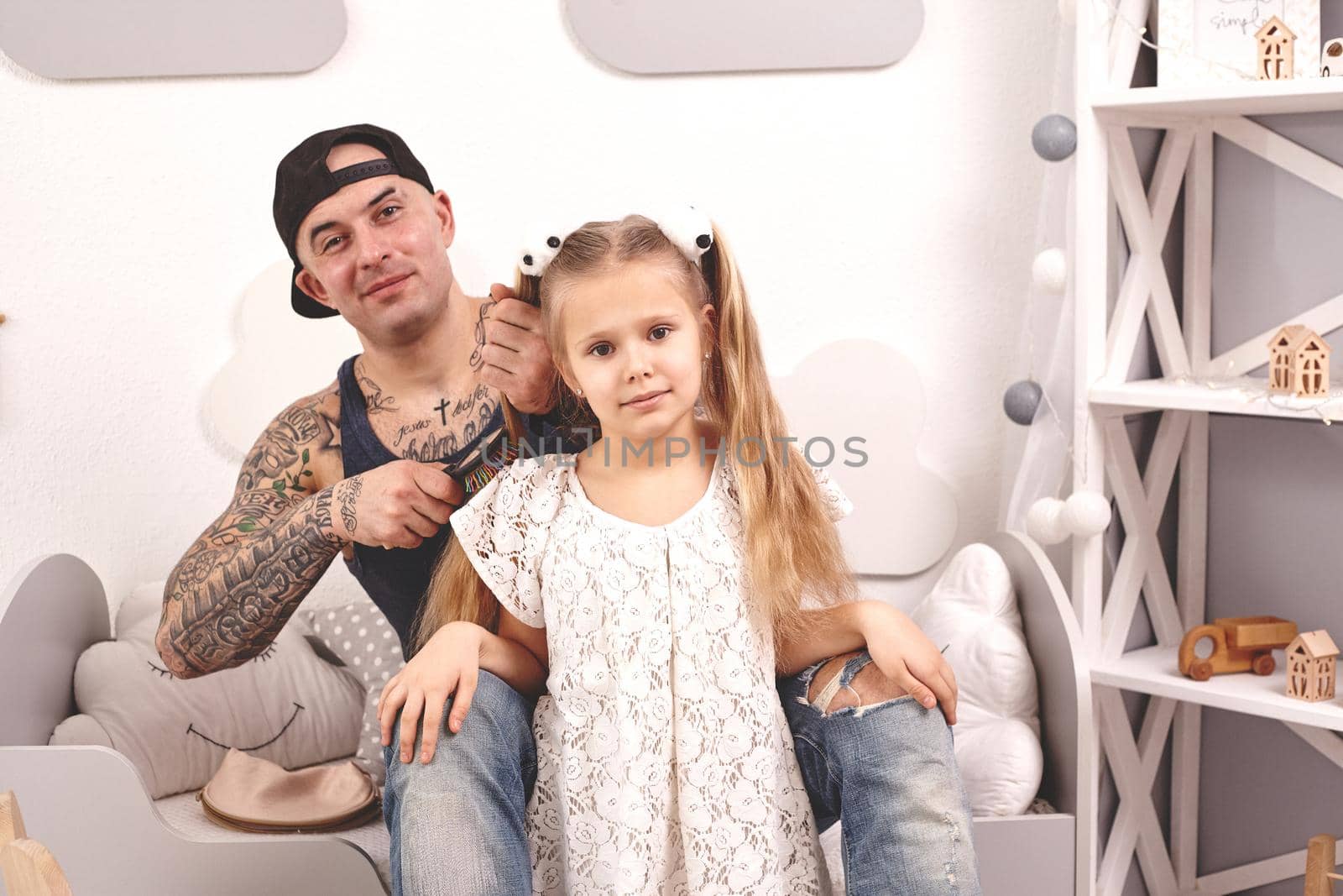 Funny time Tattoed father in a cap and his child are playing at home. Dad is doing his daughter's hair in her bedroom. They are looking happy. Family holiday and togetherness.