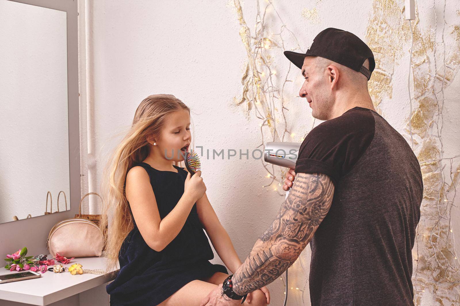 Cute little daughter and her tattoed dad are playing together near a mirror. Dad is drying his daughter's hair while she is singing in the hairbrush. Family holiday and togetherness.