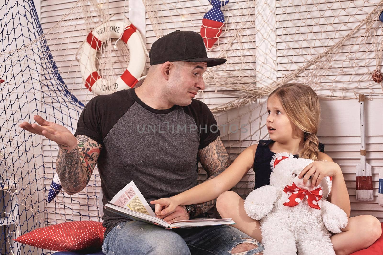 Athletic tattoed man in a cap is spending time with his little cute daughter. They are sitting on a white bench with some red pillows, in a room decorated in a marine style. She is holding a toy bear, pointing on something in a book and looking at her daddy. Reading fairytales while daughter is sitting nearby. Happy family.