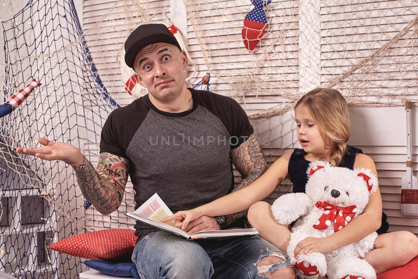 Handsome tattoed man is spending time with his little cute daughter. Reading fairytales while daughter is sitting nearby. Happy family. by nazarovsergey