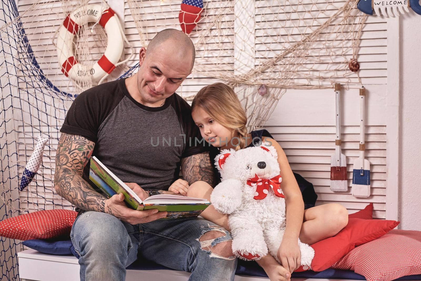 Bald tattoed man in a cap is spending time with his little cute daughter. They are sitting on a white bench with some red pillows, in a room decorated in a marine style. She is holding a toy bear. Reading fairytales while daughter is sitting nearby. Happy family.