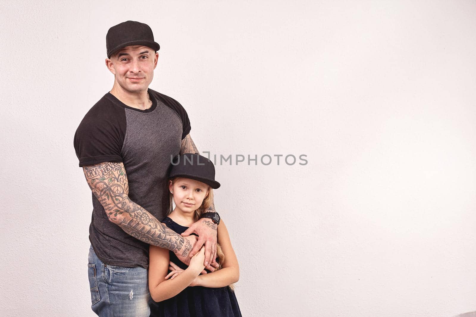 Cute little girl and her handsome tattoed father are looking at the camera and hugging, on a gray background. Each of them have a cap on their heads.