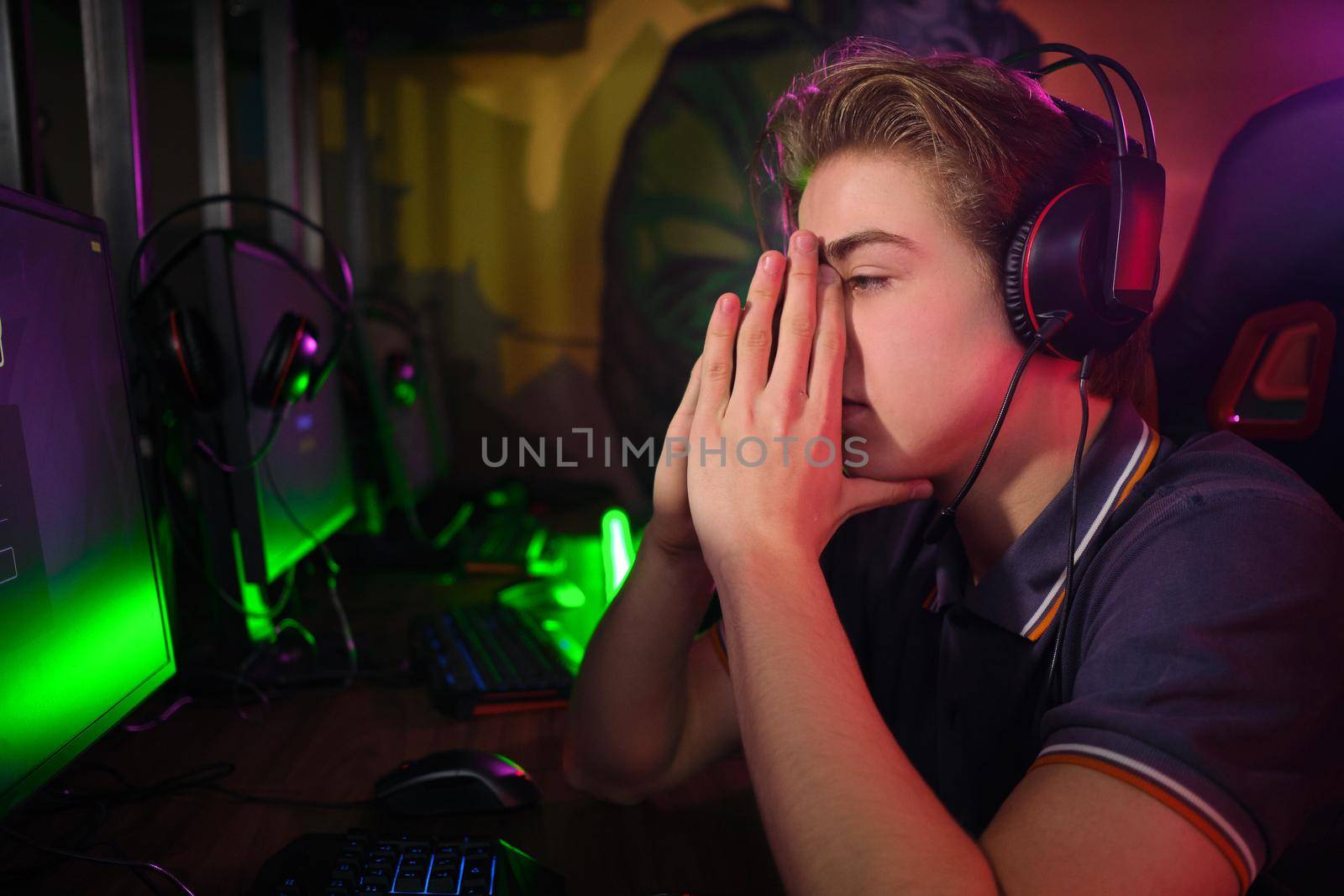 Photo of professional young caucasian gamer playing online on his PC in gaming club or cyber arena. He looks displeased and upset. Game plan went wrong
