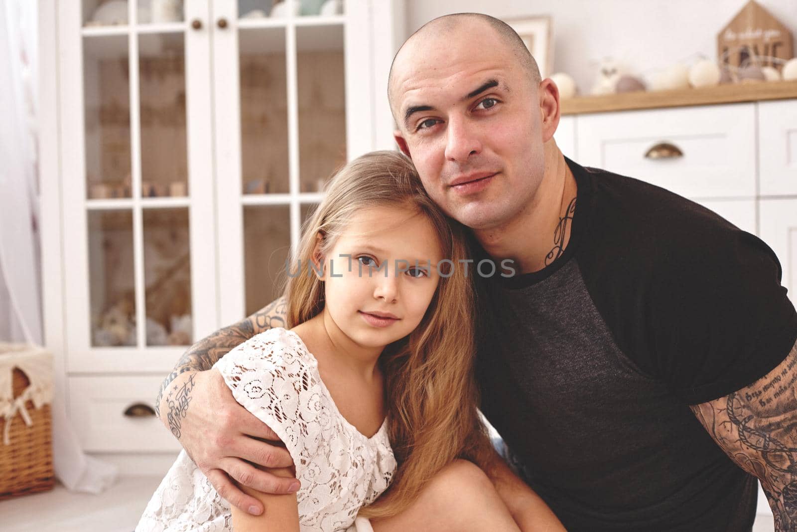 White modern kid's room whith a wooden furniture. Adorable daughter whith a long blond hair wearing a white dress . Young daddy with tattoos is hugging her and they are looking at the camera. Friendly family spending their free time together sitting on a pillows.
